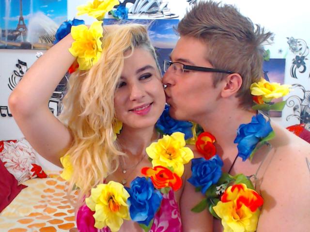 BranlePourNous, avec ses cheveux blonds et sa poitrine, portant des colliers de fleurs colorés, est embrassée sur la joue par un homme brun aux lunettes. Tous deux sourient. (photo)
