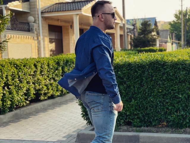 MarkusFit with a blue denim jacket, black shirt, and blue jeans, wearing sunglasses, standing on a sidewalk with green hedges in the background. His ass is slightly visible.