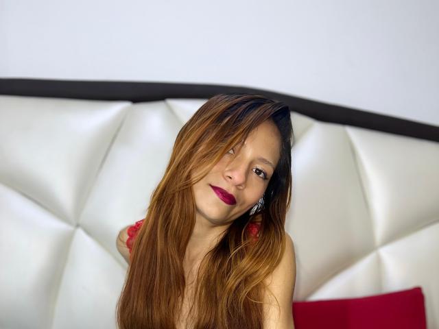 KarolCrystal with long brown hair, red lipstick, and subtle makeup, sitting on a white, padded headboard, showing off her bare shoulders and a cute, confident smile.