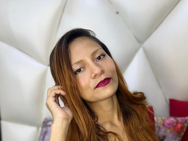 KarolCrystal with long brown hair, purple lipstick, and slight smile, touching her head with her right hand, white tufted background.