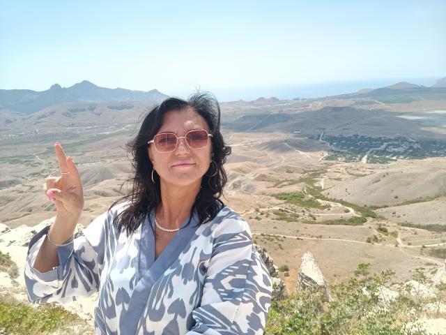 DianaLaNeon with wavy black hair, pink sunglasses, gray patterned shirt, and silver necklace, holding up a finger, smiling, on a sunny hill with a clear blue sky.