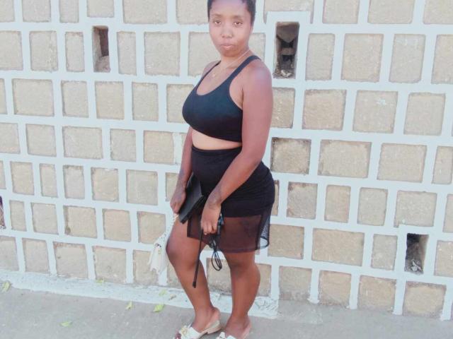 Jobba has dark brown skin, medium-sized boobs, and a curvy ass. She's wearing a black sports bra and skirt, holding a bag, with a brick wall behind her.