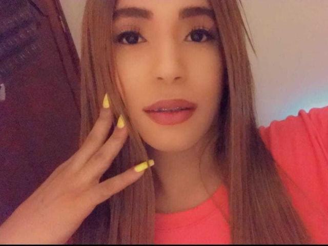 AndreaHotBigCock has long brown hair, yellow nails, pink lips, and a cute face with a slight smile. She's wearing a pink top and touching her hair.