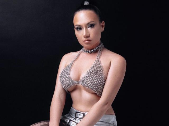 YlianaArthur con top da bikini argentato che mette in mostra il suo seno, indossa una collana con borchie e una cintura argentata, sfondo nero, capelli raccolti in una coda alta, espressione seria..