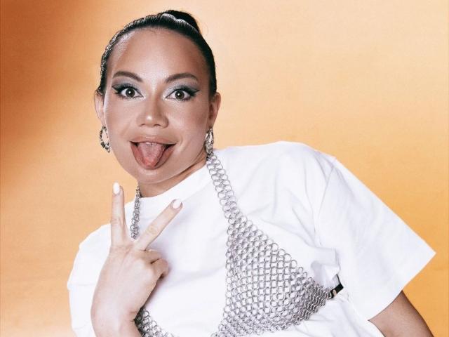 YlianaArthur mit dunkler Haut, glänzendem Make-up und hochgesteckten Haaren, trägt ein weißes Hemd mit einer silbernen Kette um den Hals, streckt ihre Zunge heraus und macht eine "rock on"-Handbewegung..