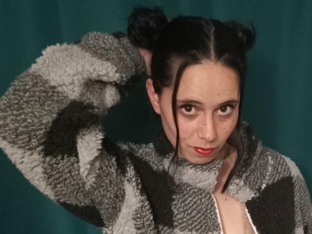 Nanastazia with dark hair in double buns, wearing a gray and black fuzzy jacket, hand in hair, red lipstick, green background, looking directly at camera.
