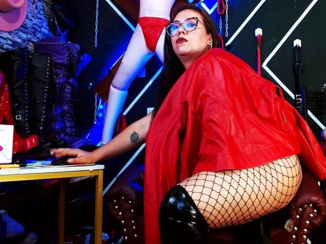 LexyDom with glasses, red jacket, fishnet pants, and black thigh-high boots, kneeling, showing her ass. Mannequin in red lingerie behind her. Tattoos visible on LexyDom's arm.