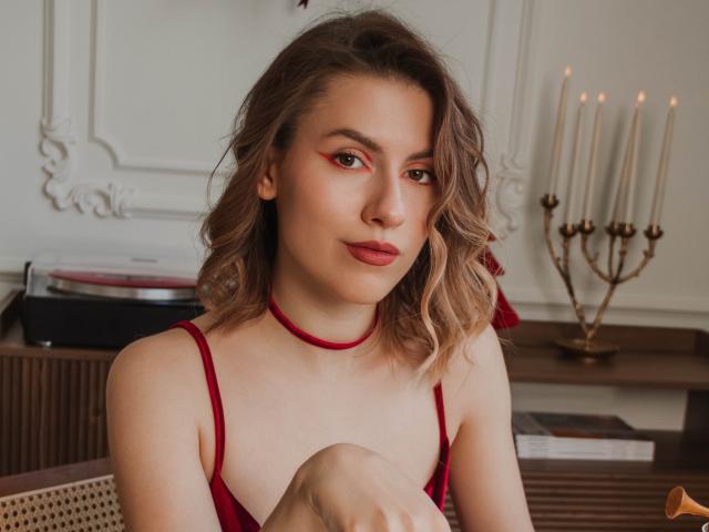 MaryBess with red lipstick, red choker, and wavy blonde hair, wearing a red velvet dress, sitting with knees up, looking at camera, candleholder in background.