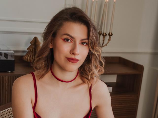 MaryBess has wavy brown hair, red eyeliner, red lipstick, and a red choker with matching red bra straps. She's smiling slightly, showing off her tits. Candleholder in background.