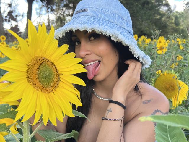 Stefeney Collin, com cabelos longos e pretos, língua de fora, lambendo uma grande girassol amarela, usando um boné azul, sem camisa, com tatuagens no braço e uma pulseira preta..