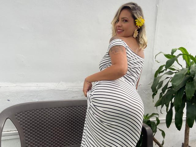 ChantalSex69, with yellow flower in hair, smiling, wearing striped dress, pulling up dress to show her curvy ass, tattoo on upper arm, standing next to chair, plant in background.