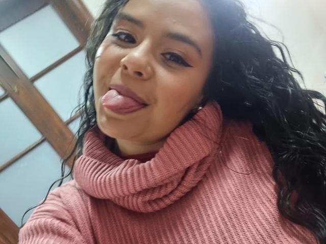 ArielleWilde with long, curly black hair, sticking out her tongue, wearing a pink rib-knit turtleneck, has bold eyeliner, and a cute, playful expression.