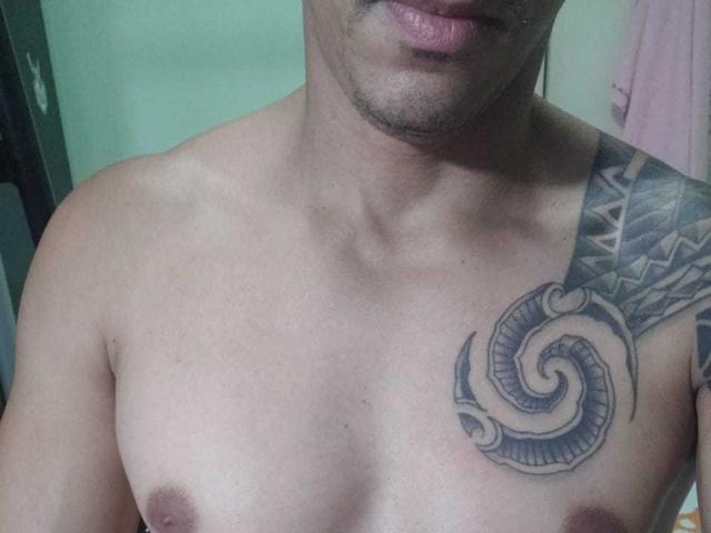 O peito de MagrinHodot está descoberto, com uma tatuagem no ombro direito, que apresenta um desenho em espiral, e os mamilos são visíveis. Sua pele é de cor marrom clara e ele tem uma pequena barba..