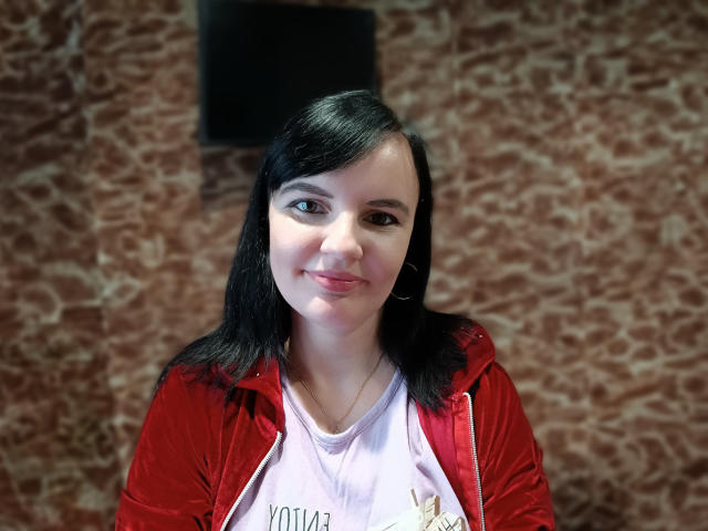 AlicexB has black hair, light skin, and small tits. She's smiling, wearing a red velvet jacket and pink shirt, with a hoop earring. She's standing in front of a brown textured wall.
