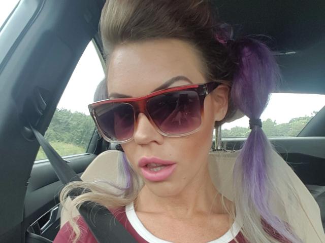Lara Bergmann – attraktiv mit lila Zöpfen, roten Sonnenbrillen, rosafarbenen Lippen und einem braunen Sicherheitsgurt, blickt im Auto nach vorne. Ihre Brüste sind teilweise unter ihrem roten Hemd sichtbar..