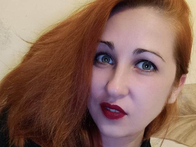 JillSilvies has pale skin, red hair, blue eyes, and red lipstick, with her hair partially covering one side of her face. She's looking directly at the camera.