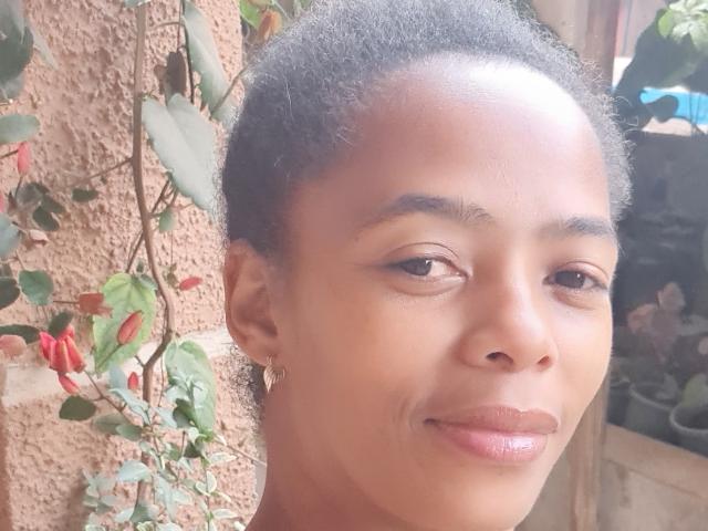ClaraElie has dark brown skin, short curly black hair, and is smiling with shiny pink lips. Her brown eyes are relaxed. She's wearing small earrings.