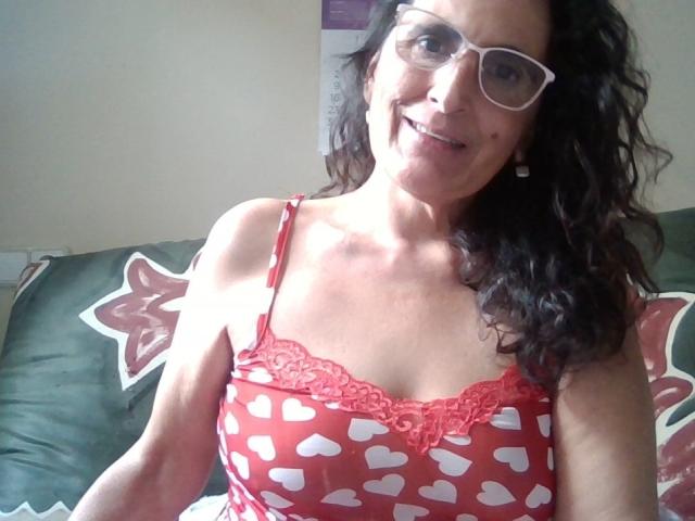 Esencia with curly black hair, glasses, and red heart-patterned camisole, smiling at the camera, showing her small tits with lace trim. She's sitting on a green patterned couch.
