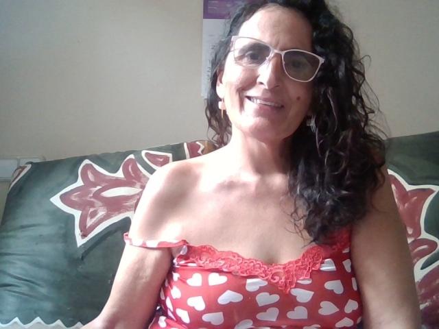 Esencia, with curly dark hair and glasses, wears a red, heart-patterned, lace-trimmed top, showing her small, perky tits and smiling. She's sitting on a green couch.