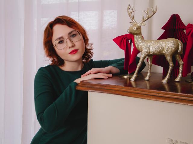 TeresaBirt with red hair, glasses, green sweater, and red lipstick, leaning on a wooden table with a gold reindeer statue and red draped objects.