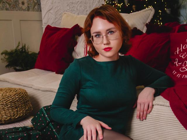 TeresaBirt with red hair, glasses, and red lipstick, wearing a green dress, sitting on a couch with red and white pillows, hands on her thighs, looking at the camera.