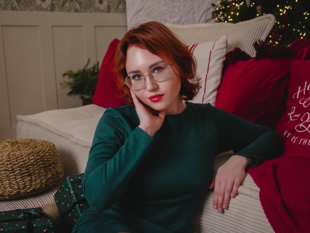 Teresa Birt, con cabello rojo, gafas, suéter verde, lápiz labial rojo, descansando en un sofá blanco, con la mano en la mejilla, almohadas rojas y verdes, cesta de mimbre, árbol de Navidad en el fondo..