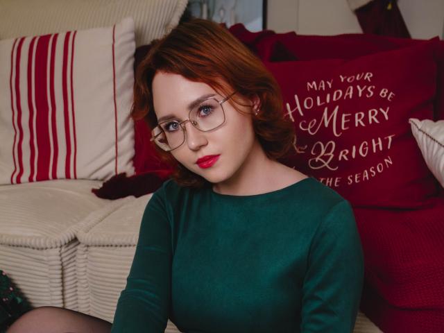 TeresaBirt with red hair, glasses, green sweater, red lipstick, sitting on couch, looking at camera, medium breasts, fair skin, Christmas decor in background, soft light.