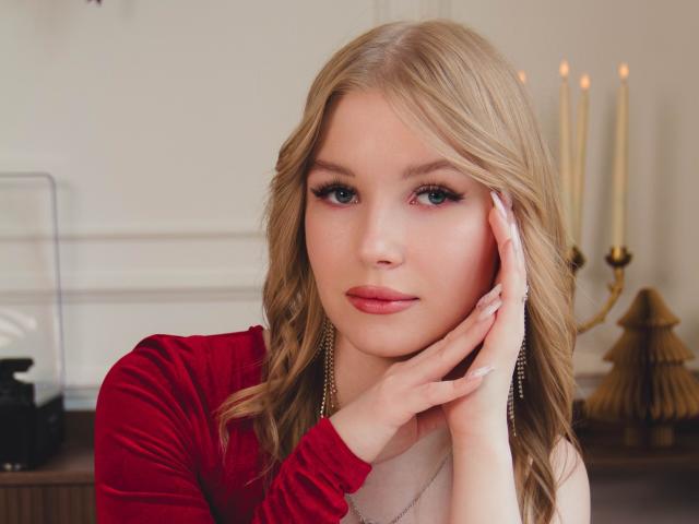 Nicole Brian hat blondes, welliges Haar, grüne Augen und trägt ein rotes Samintop mit silbernen Ohrringen. Sie berührt ihre Wange mit einer Hand, blickt mit einem leichten Lächeln in die Kamera..