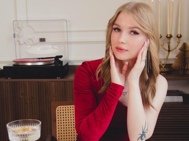 NicoleBrian with blonde hair, red velvet dress, hand on cheek, earrings, tattoo on right arm, looking at camera, table with glass, candles, and vintage record player in background.