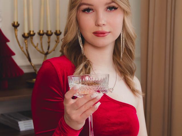 NicoleBrian with blonde hair, red velvet dress, and gold earrings, holding a glass of pink champagne, looking at the camera with a slight smile. Boobs, tits, pussy, ass not visible.