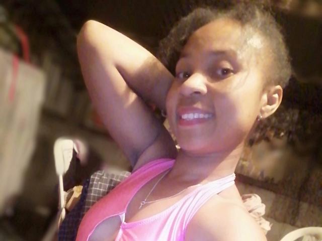Edrwina69 has dark brown skin, small round tits, and a pink halter top, smiling with her arm behind her head, showing curly hair and a cute face.