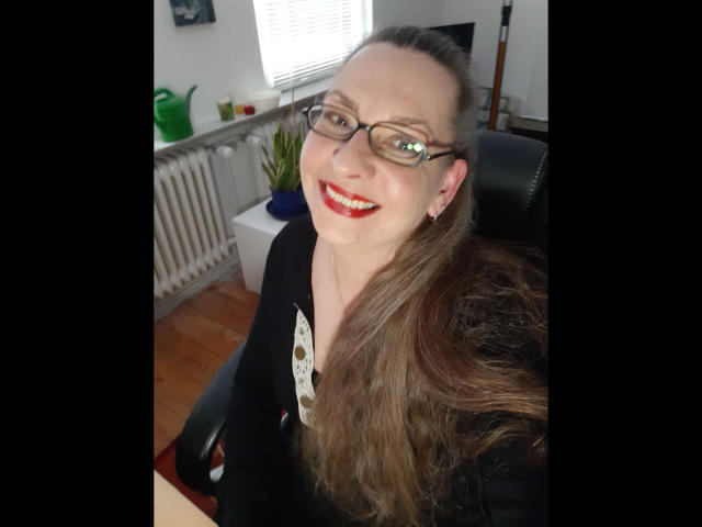 CashLadyVivian: una donna attraente, con lunghi capelli castani, occhiali, rossetto rosso e una maglia nera, che sorride alla fotocamera. Ha il seno piccolo e indossa una camicia bianca con bottoni..