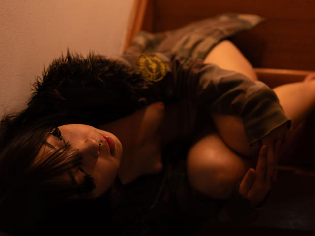 MillaHottLove69 with dark hair, wearing a fur-trimmed jacket, has her tits pressed against a wooden surface, looking up with a slightly open mouth, in a dimly lit room.
