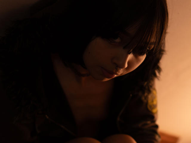 MillaHottLove69 has dark, messy hair, a nose ring, and a serious, slightly curious expression in dim, warm light. She's wearing a dark, fuzzy jacket.