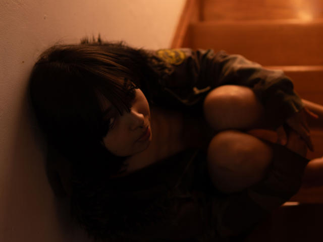 MillaHottLove69 is crouched on wooden stairs, wearing a dark, patterned jacket, with messy black hair covering her eyes, in low, moody lighting.