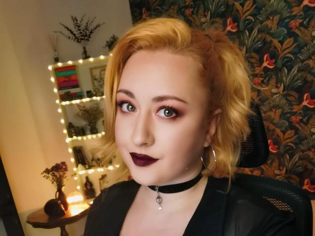 LuxNoir has blonde puffy hair, dark purple lipstick, hoop earrings, black choker with pendant, and is wearing a black top. Her makeup is smoky with green eyes.