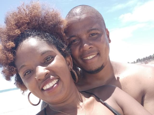 TropicalCouple: Eine dunkelhäutige Frau mit lockigem, braunem Haar, Reifenohrringen und einem Nasenring, und ein dunkelhäutiger Mann mit rasierter Kopfhaut, der in die Kamera lächelt..
