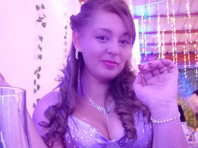 AmandaHotL with purple-tinted skin, curly brown hair, and a sparkly blue dress showing her big tits, wearing a diamond necklace, holding a glass, smiling.