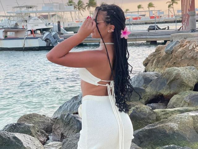AlessiaFerry con lunghe trecce nere, top da bikini bianco che mostra i suoi piccoli seni, gonna bianca, fiore rosa, che si copre la bocca, in piedi vicino all'acqua rocciosa, con un porto sullo sfondo..