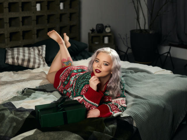 EnergyBurnny with purple hair, red sweater, and white skin, lying on bed, legs up, holding green box, tattoo on right thigh, red lipstick, and smiling.