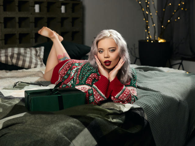 EnergyBurnny with silver hair, red lipstick, green and red sweater, lying on bed, hands on cheeks, legs up, green box in front, dark red panties visible, tattoo on thigh.
