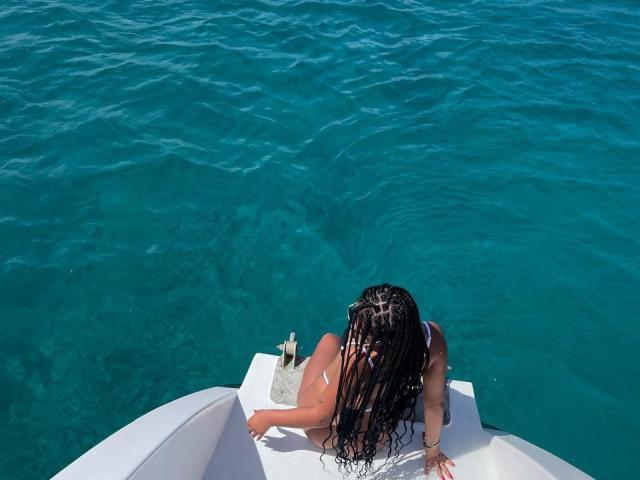 AlessiaFerry with long braided hair, wearing a white bikini, sitting on a white boat, back to camera, facing bright turquoise water, small white bag on boat.