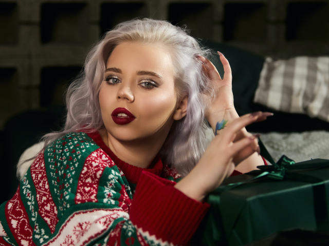EnergyBurnny with light purple hair, red lipstick, and green-red Christmas sweater, hand raised, looking at camera, gift box with green ribbon in foreground, dark background.