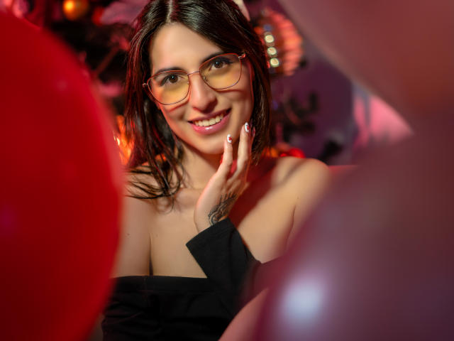 IsaDupont with glasses, smiling, black off-shoulder top, hand touching face, red and pink balloons in foreground, medium-length brown hair, cute, sexy, bokeh effect, indoor lighting.