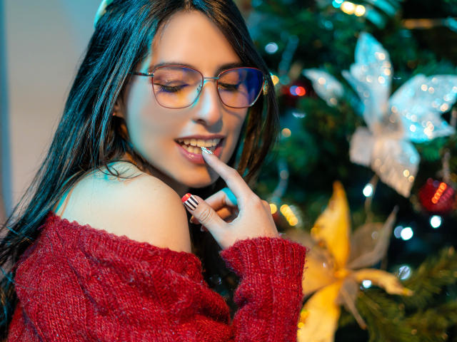 IsaDupont with glasses, red off-shoulder sweater, finger near her slightly open mouth, showing her teeth, and dark wet hair, standing next to a decorated Christmas tree.