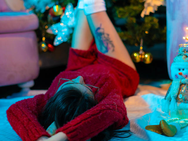 IsaDupont lying on her back with legs up, wearing a red sweater, no pants, showing a tattoo on her thigh, black hair, red glasses, white socks. Christmas lights.
