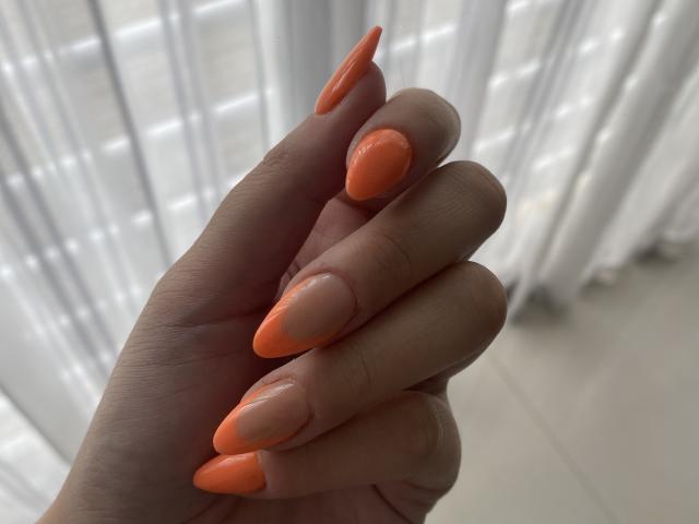 AlexaLip's hand with long, sharp, neon orange nails on her fingers, showcasing a glossy, nude-painted thumb against a soft, white, blurred background.