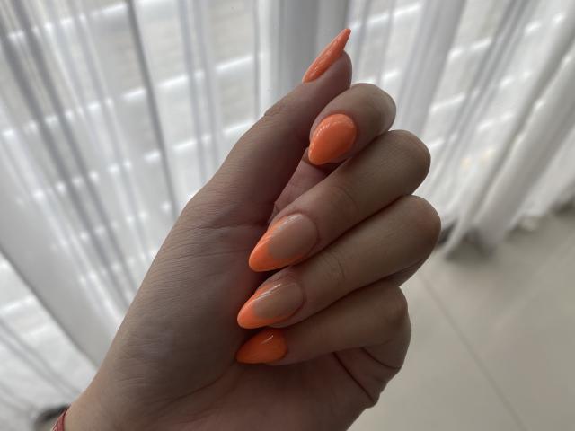 AlexaLip's hand with shiny, orange, almond-shaped nails, fingers slightly curled, light skin, and soft texture, against a blurred white curtain background.
