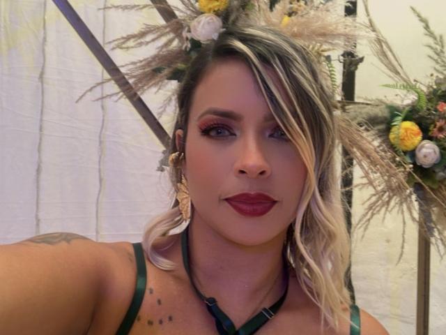 ChantalSex69 with blonde hair, red lipstick, green straps, and flower headpiece, showing tattoos on shoulder, wearing gold earrings, looking at camera with confident smile.