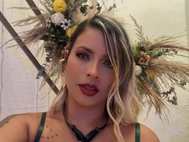 ChantalSex69 tiene cabello rubio, lápiz labial rojo y tirantes verdes, y lleva un tocado floral con flores amarillas y blancas. Sus tatuajes son visibles en su pecho..